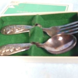 May include: A set of silver baby cutlery in a green and white box. The set includes a spoon and fork with floral designs.