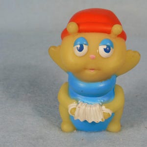 Puede incluir: Un juguete de goma amarillo con un sombrero rojo, una bufanda azul y un acordeón blanco. El juguete tiene una cara amigable con ojos azules y una nariz rosa.