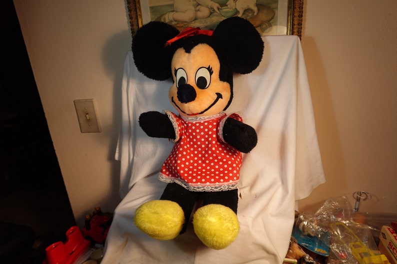 May include: A large plush Minnie Mouse toy with black fur, red polka dot dress, and yellow shoes. The toy is sitting on a white surface.