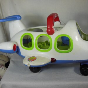 May include: A white toy airplane with a blue base, tail, and engine accents. It features three bright green window frames, a red handle on top, and black wheels. The side wing displays a circular sticker with a colorful palm tree graphic design.