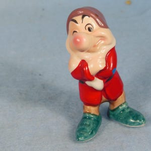 May include: A ceramic figurine of a dwarf character, with a red outfit, green shoes, and a brown cap. The figurine has a rosy complexion and is posed with arms crossed, winking. The figure is a collectible item.