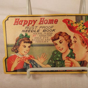 Könnte beinhalten: Vintage Happy Home Rostfreier Nadelkasten mit 85 sortierten Goldaugennadeln und einem Nadelfadenzieher. Das Buch zeigt eine farbenfrohe Illustration von zwei Frauen beim Nähen.