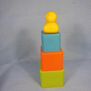 Peut inclure: Une pile de quatre blocs colorés, avec un canard jaune sur le dessus. Les blocs sont vert, orange, bleu et jaune.
