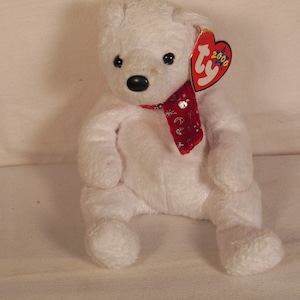 Puede incluir: Oso polar de peluche blanco con una bufanda roja y blanca con un diseño de copo de nieve plateado. El oso tiene ojos negros y una nariz negra. El oso está sentado sobre una superficie blanca. El oso tiene una etiqueta Ty pegada a su oreja que dice "Ty 2000".