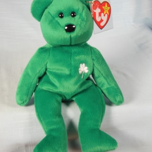 May include: Green plush teddy bear with a black nose and a white shamrock on its chest. The bear is sitting with its legs crossed and has a Ty tag attached to its ear.