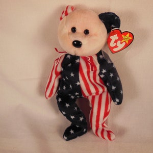 May include: A plush toy bear wearing a red, white, and blue outfit with stars and stripes. The bear has a black nose and brown eyes. The bear is wearing a Ty tag that says "Beanie Babies".