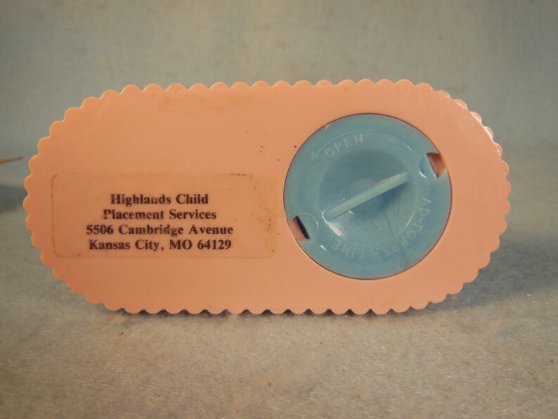 May include: Pink plastic piggy bank with a blue lid that says "OPEN" and "INSERT COIN". The bottom of the bank has the text "Highlands Child Placement Services 5506 Cambridge Avenue Kansas City, MO 64129".