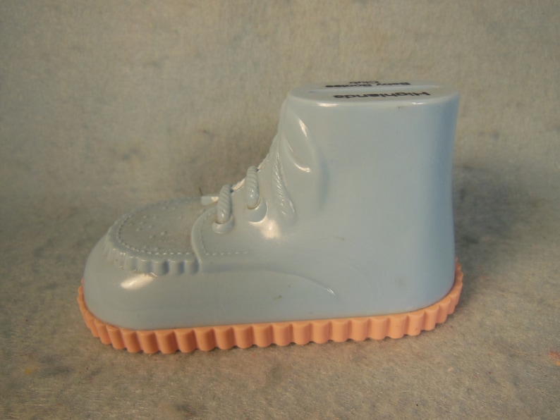 May include: A light blue plastic baby shoe shaped piggy bank with a pink bottom and the text "Highchair" on the side.