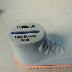 May include: A blue plastic baby shoe piggy bank with a slot on top. The shoe has a pink bottom and says "Highlands Baby Bootee Club" on the side.