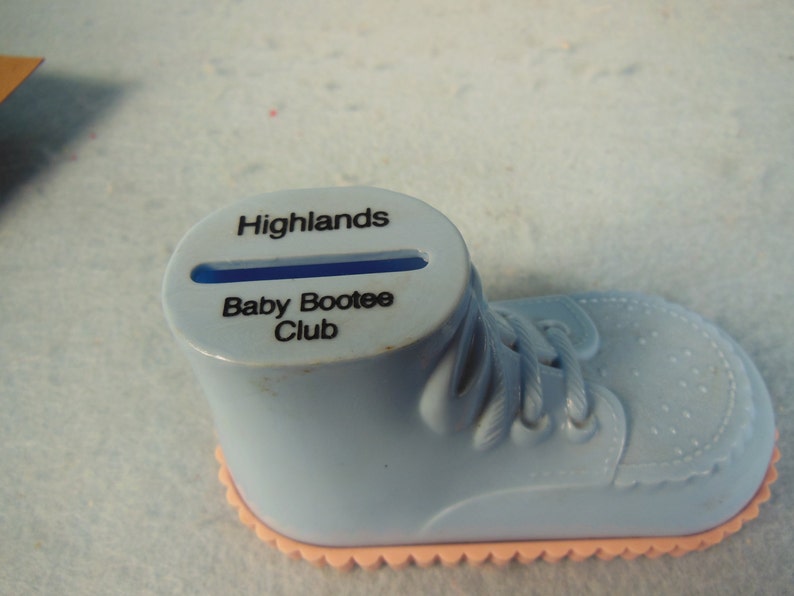 May include: A blue plastic baby shoe piggy bank with a slot on top. The shoe has a pink bottom and says "Highlands Baby Bootee Club" on the side.