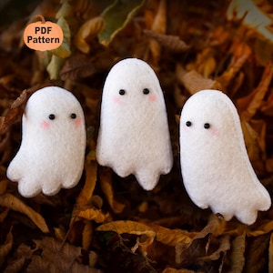 May include: Three white felt ghost decorations with black eyes and pink cheeks are arranged on a bed of autumn leaves. The ghosts have a simple, friendly design. A pumpkin-shaped graphic with the text "PDF Pattern" is in the upper left corner.