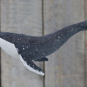May include: A gray felt whale with white and black bead accents hanging from a string. The whale is a handmade toy.