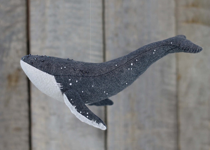 May include: A gray felt whale with white and black bead accents hanging from a string. The whale is a handmade toy.