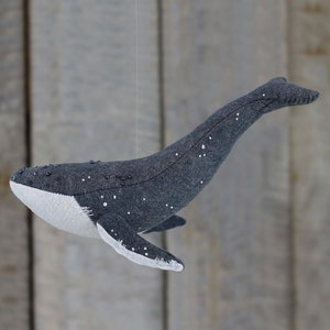 May include: A gray felt whale with white felt accents and black beads sewn on. The whale is hanging from a string.