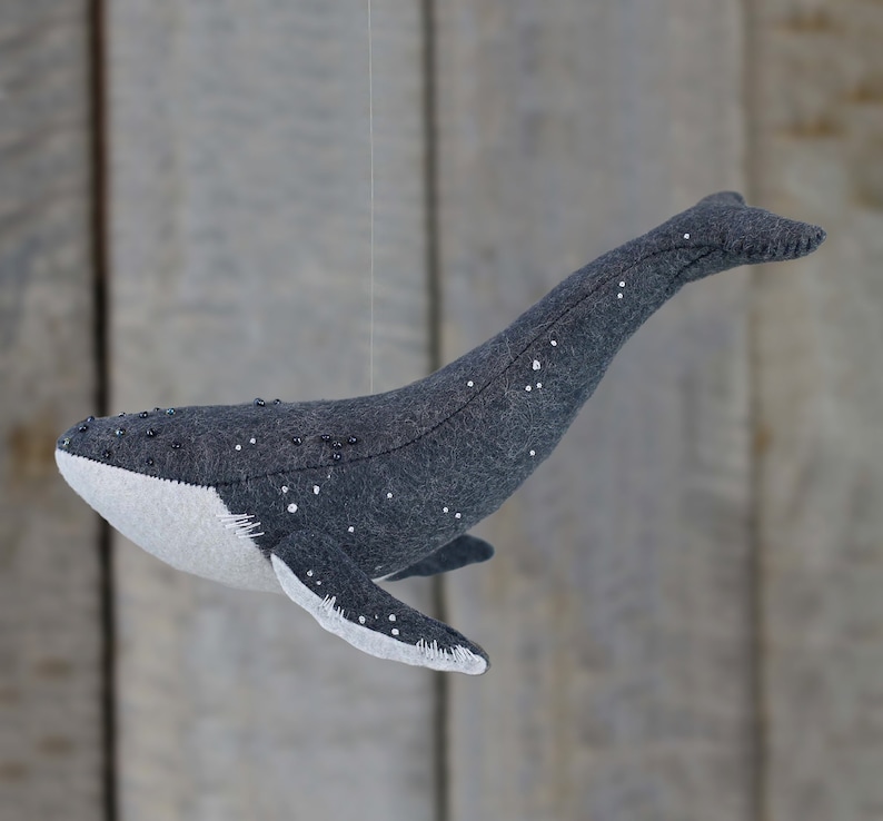 May include: A gray felt whale with white felt accents and black beads sewn on. The whale is hanging from a string.