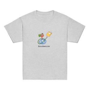 Könnte beinhalten: Ein heidefarbenes T-Shirt mit einem Cartoon-Wurm, der einen grünen Hut trägt und ein Buch hält. Der Text "Bookworm" ist unter dem Bild gedruckt.