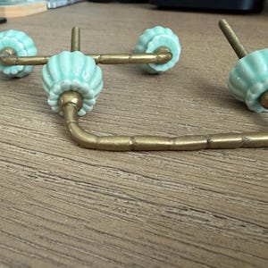 May include: Two drawer pulls featuring a curved, antique brass-toned metal handle with a bamboo-like texture. Each handle is attached to two fluted, pale mint green ceramic knobs that have a scalloped edge and a threaded metal bolt for mounting.