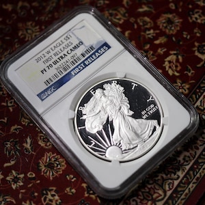 May include: A silver coin in a protective case. The coin features a detailed depiction of a woman and the text "IN GOD WE TRUST". The case has a label with the text "2012 W EAGLE S$1 FIRST RELEASES PF 70 ULTRA CAMEO".