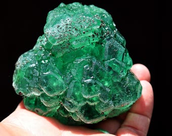 May include: A vibrant green fluorite crystal cluster featuring translucent, geometric cubic formations with small, dark metallic mineral inclusions scattered across the textured, multifaceted surface of the raw mineral specimen.