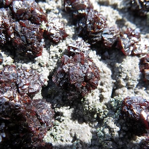 Può includere: Primo piano di un gruppo di cristalli rosso scuro su una superficie grigia e testurizzata. I cristalli sono affilati e hanno un bagliore metallico.