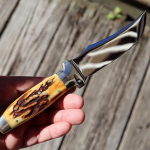 May include: A hunting knife with a curved, polished steel blade and a handle featuring a cream and brown pattern. The knife has a silver bolster and a riveted handle. The blade reflects the surrounding environment.