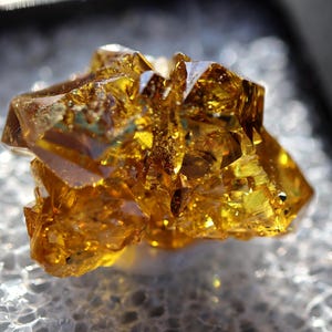 May include: A translucent, golden-yellow mineral specimen featuring sharp, angular crystalline structures with a high-gloss, reflective surface. The cluster displays deep amber hues and complex geometric facets that catch the light, creating a brilliant, multi-dimensional appearance throughout the entire formation.