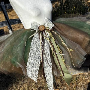 May include: A layered tulle tutu skirt in shades of forest green and brown, featuring a decorative front accent. The embellishment includes a white floral lace bow, a central dark brown wooden button, and hanging ribbons in brown and olive green.