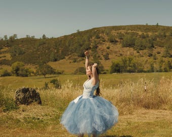 May include: A light blue dress featuring a fitted bodice and a voluminous, multi-layered tulle skirt that reaches mid-calf. The garment has a sleeveless design with a V-neckline and is paired with light-colored, strappy heeled sandals.