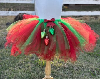 May include: A layered tulle tutu skirt in alternating red and green colors, featuring a dark red ribbon bow at the center waist. Three glittery, bulb-shaped ornaments in red and green hang from the bow, resting against the skirt's fabric.