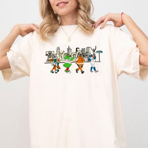 May include: Cream-coloured t-shirt with a colourful graphic of cartoon characters walking in front of a city skyline. The word "LOVE" is incorporated into the design. The characters are wearing sports uniforms.