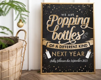 May include: A black chalkboard sign with gold lettering announcing "We are popping bottles of a different kind next year." The sign also includes the text "Baby Johnson due September 2023" and two gold bottle images.