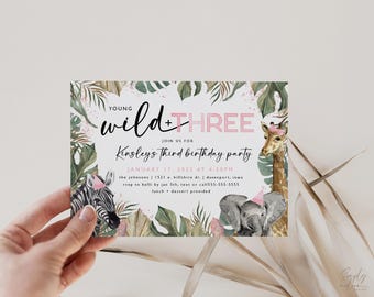 Puede incluir: Una invitación de cumpleaños con el texto "Young Wild + Three". La invitación presenta ilustraciones de una cebra, una jirafa y un elefante, todos con sombreros de fiesta, rodeados de hojas tropicales y acentos rosas. La invitación es sostenida por una mano.