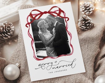 May include: A white card with a black and white photo of a couple framed by a red watercolor bow and ribbon. The text "merry + married" is written in cursive, with "THE JOHNSONS" below.