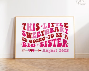 May include: A framed print with the text "THIS LITTLE SWEETHEART IS GOING TO BE A BIG SISTER August 2025" in shades of pink and red. The design includes hearts and an arrow. The frame is gold.