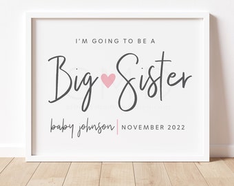 Op de afbeelding: Een witte ingelijste print met zwarte tekst die "I'm going to be a Big Sister baby johnson NOVEMBER 2022" luidt. Een roze hart bevindt zich tussen de woorden "Big" en "Sister".