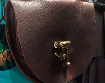 May include: A brown leather bag with a brass clasp. The bag is attached to a brown leather belt with a buckle.
