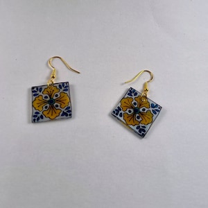 May include: A pair of gold dangle earrings with square-shaped blue and white ceramic tiles featuring a yellow floral design.
