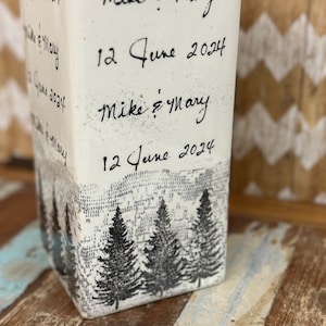 May include: White ceramic vase with a rustic design featuring a forest scene and the text "Mike & Mary 12 June 2024" repeated twice.