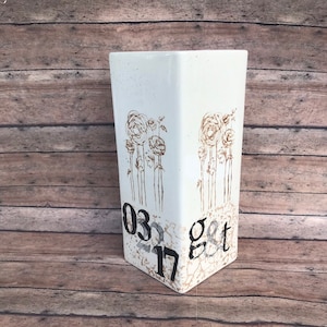 May include: White ceramic vase with a distressed finish and a floral design. The vase has the numbers 03 17 and the text "g&t" printed on it.
