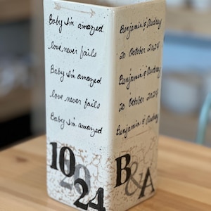 May include: White ceramic vase with black handwritten text that reads "Baby I'm amazed. Love never fails. Benjamin & Audrey. 20 October 2024." The vase also has black numbers 10, 20, and 24, and the letters B & A.
