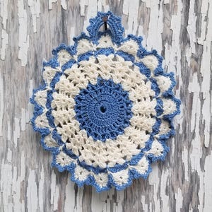 May include: A round, hand-crocheted potholder featuring a central blue circle surrounded by alternating rings of cream and blue yarn. The design includes a scalloped edge and a small loop at the top for hanging, showcasing a detailed, textured stitch pattern.