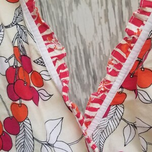 May include: Cream-colored fabric with a cherry pattern in shades of red and orange, accented with black and white leaves. The edges are trimmed with a red and white striped ruffle. A summer-themed garment.