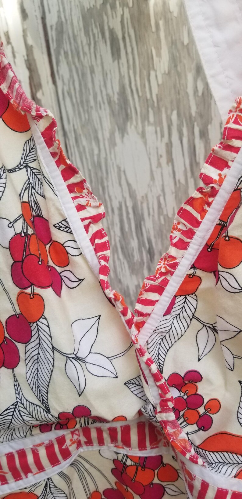 May include: Cream-colored fabric with a cherry pattern in shades of red and orange, accented with black and white leaves. The edges are trimmed with a red and white striped ruffle. A summer-themed garment.