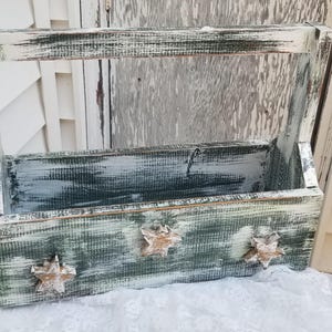 May include: A rectangular wooden caddy with a tall handle, featuring a distressed finish in shades of dark green and white. The front panel is decorated with three small, light-colored wooden star cutouts spaced evenly across the surface.