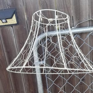 May include: A white wire lampshade frame featuring a hexagonal mesh pattern. The structure consists of a circular top ring, a wider circular bottom ring, and vertical support struts connecting the two, with a central fitter at the top.