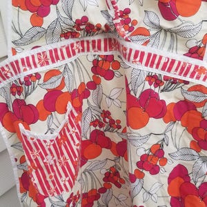May include: Apron with a cream background and a vibrant cherry pattern in shades of red and orange. The apron features a pocket with red and white stripes and white trim.
