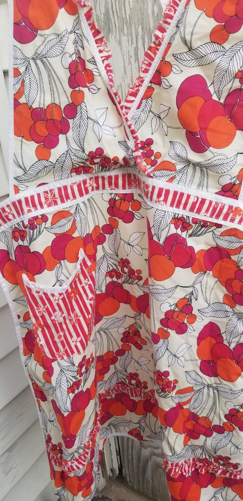 May include: Apron with a cream background and a vibrant cherry pattern in shades of red and orange. The apron features a pocket with red and white stripes and white trim.