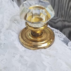 May include: A door knob featuring a clear, faceted glass handle attached to a circular, polished brass base. The metal base shows signs of wear and patina, while the glass top has a multi-sided geometric shape that reflects light.