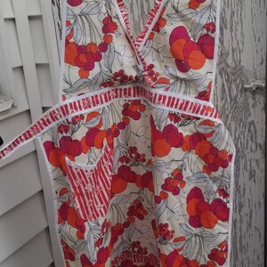 May include: A vintage apron with a cream background and a vibrant floral pattern of red and orange fruit. The apron has a V-neck, a pocket with red and white stripes, and white trim. It is hanging on a black hanger.