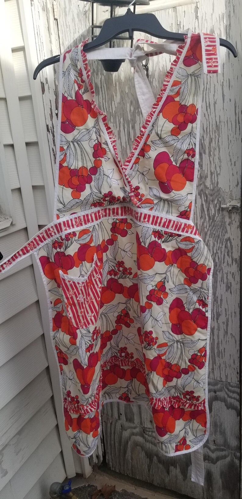 May include: A vintage apron with a cream background and a vibrant floral pattern of red and orange fruit. The apron has a V-neck, a pocket with red and white stripes, and white trim. It is hanging on a black hanger.